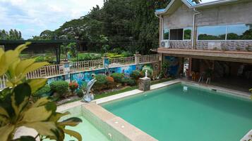 A swimming pool area with a house in the background, surrounded by greenery and a decorative fence.