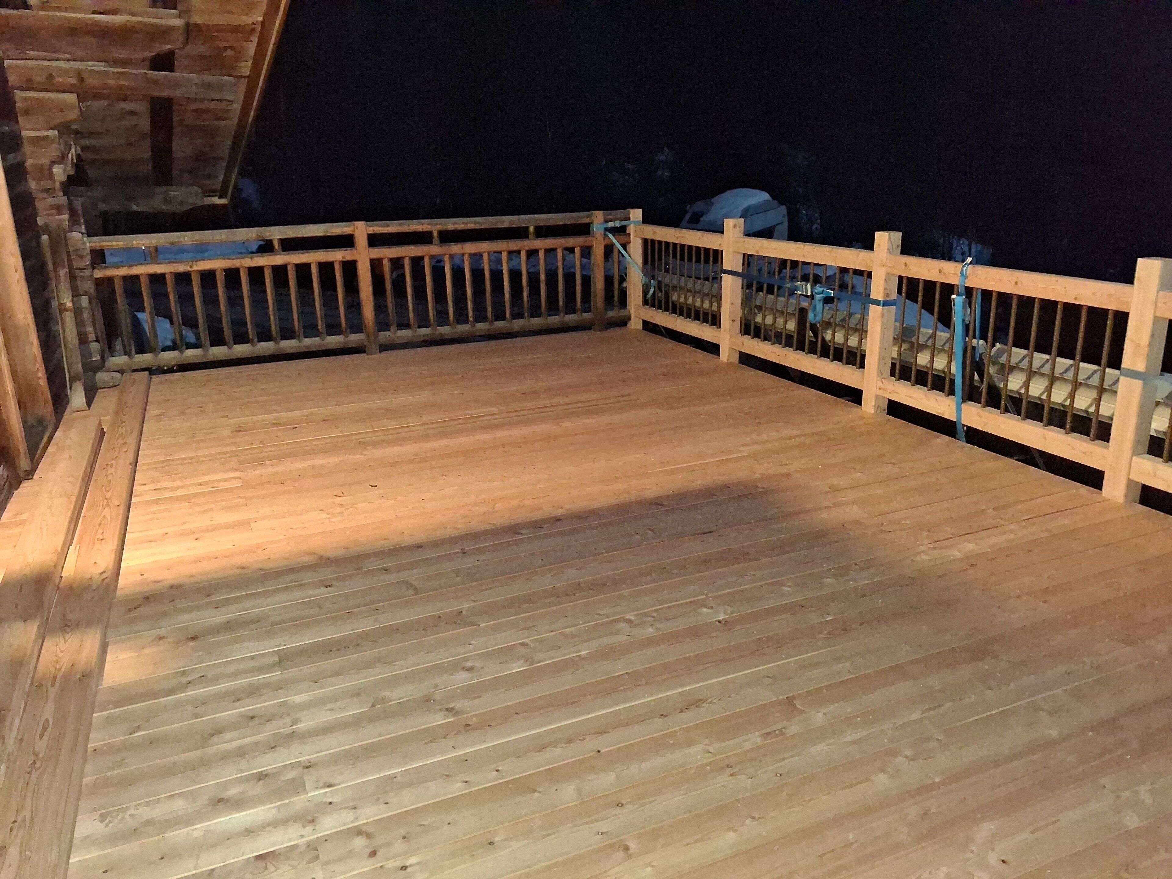Une terrasse en bois avec une rambarde, un mur en pierre et un véhicule à l’arrière-plan.