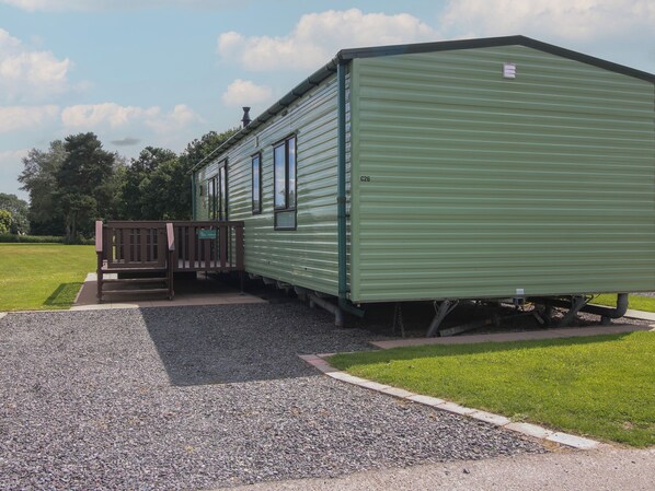 A green mobile home with a porch and a small deck, situated in a grassy area with trees in the background. - Mallard (Leominster)