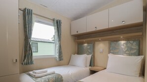 A twin room with two beds, a window with curtains, a wooden headboard, and a small bedside table. - Mallard (Leominster)