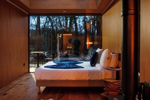 A modern bedroom with a large bed, wooden flooring, and a view of the outdoors.