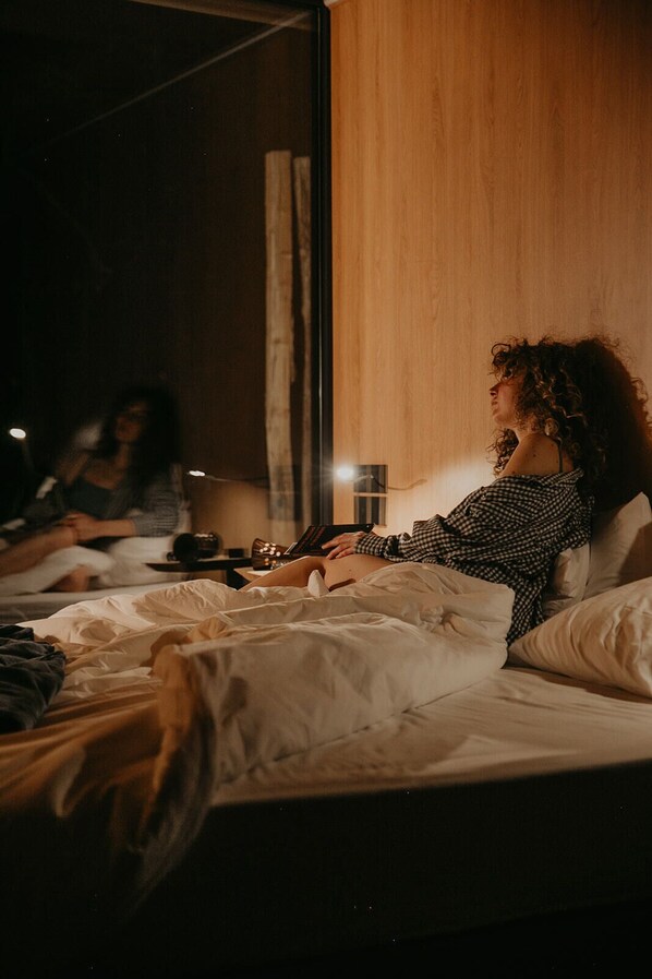 A person lying in bed with a laptop, a bedside table with a lamp, and a window with curtains.