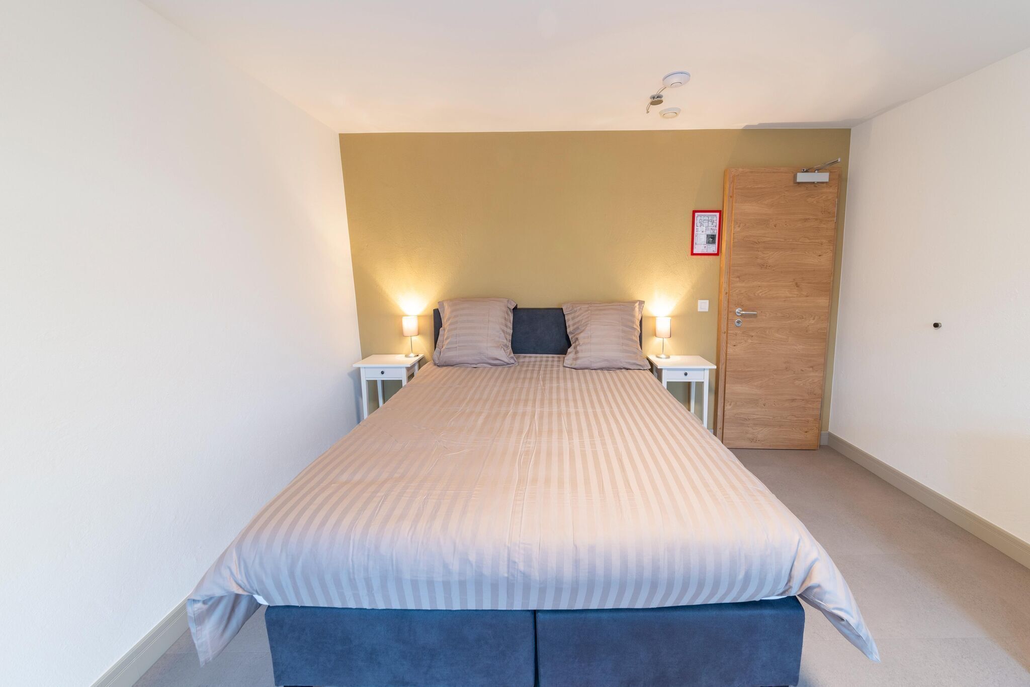 Une chambre à coucher comprenant un lit, deux tables de chevet, une commode, une porte en bois et un luminaire au plafond.