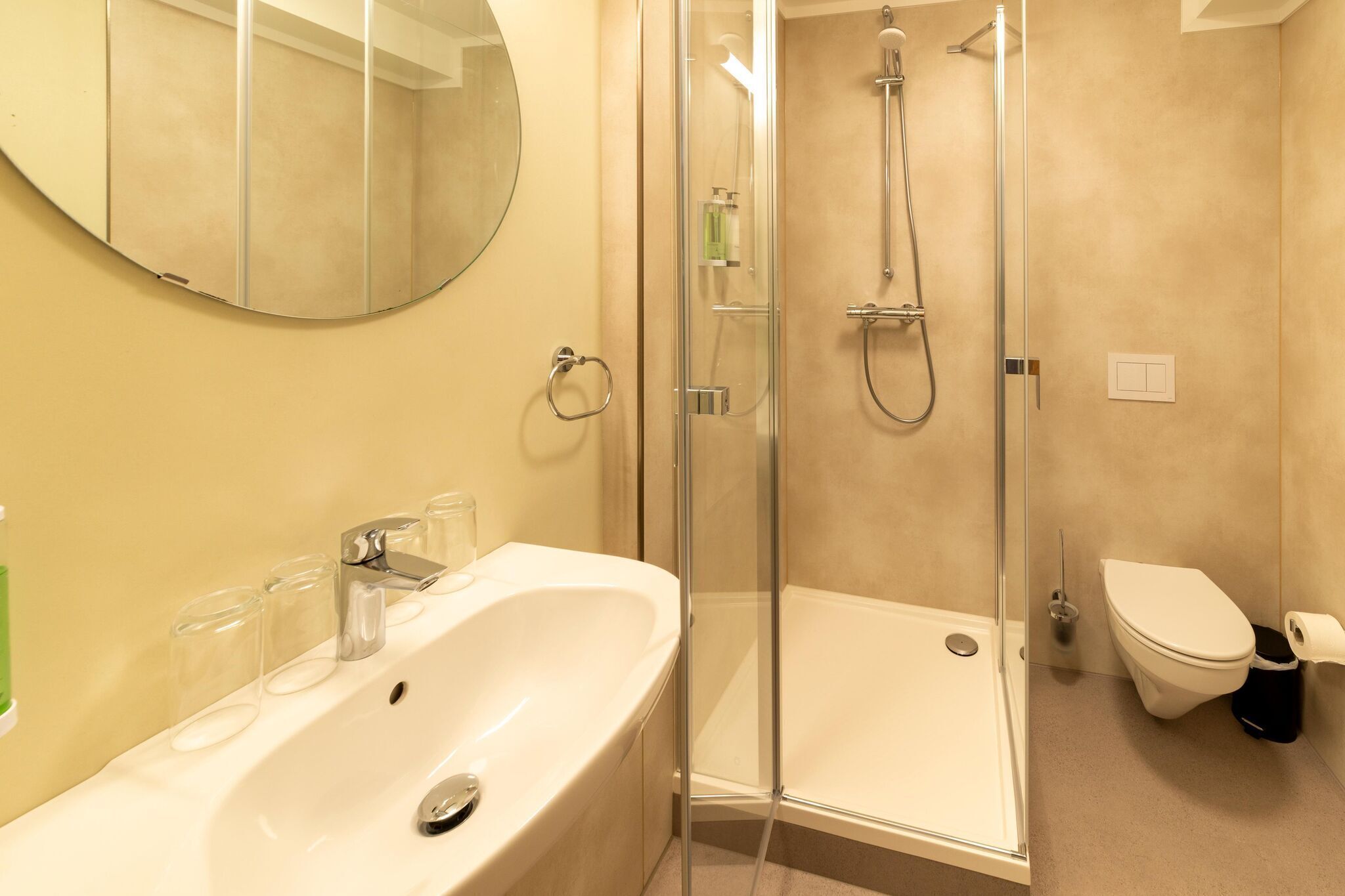 Een moderne badkamer met een douchecabine van glas, een witte wastafel met glazen bekers en een toilet.