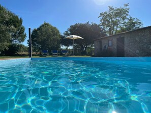 Une piscine aux eaux cristallines et bleues, entourée d’arbres et bordée d’un bâtiment en pierre.