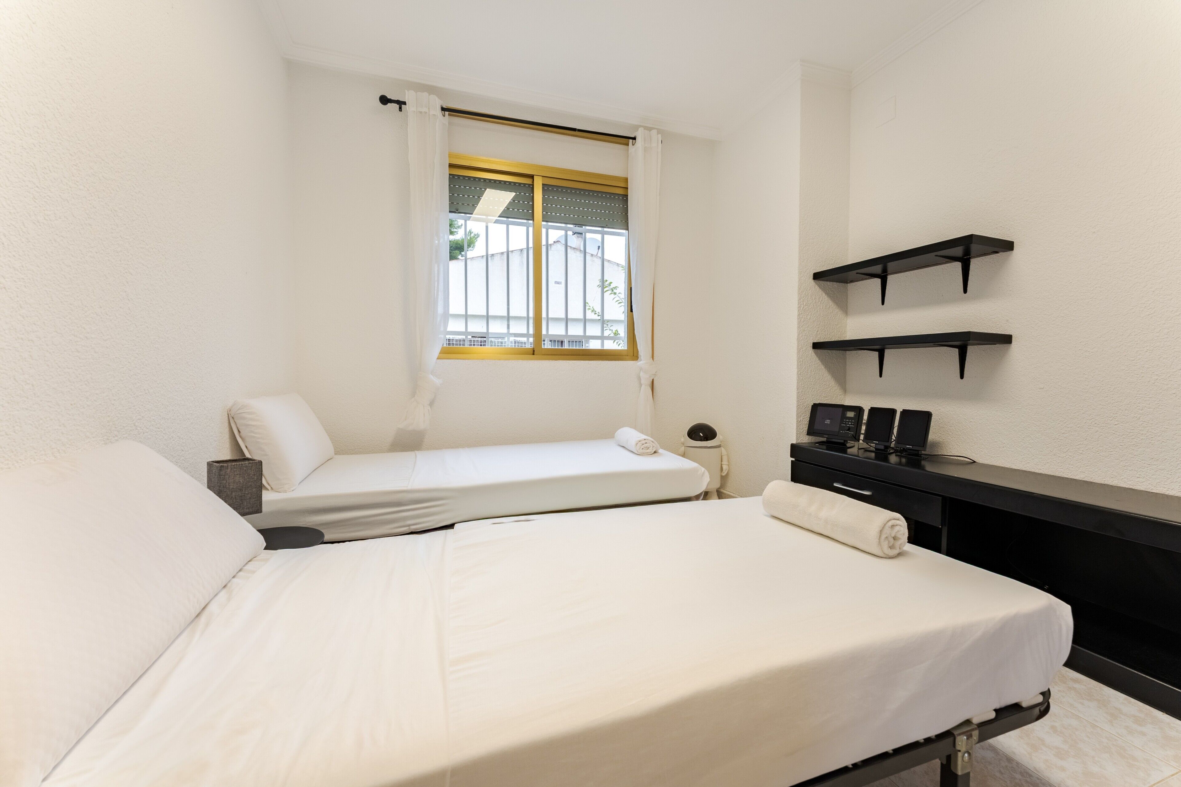 Een hotelkamer met twee bedden, een raam met gordijnen, een bureau met stoel, en twee wandplanken.