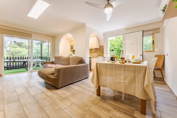 A spacious living area with a sofa, dining table, and a view of a garden through large glass doors.