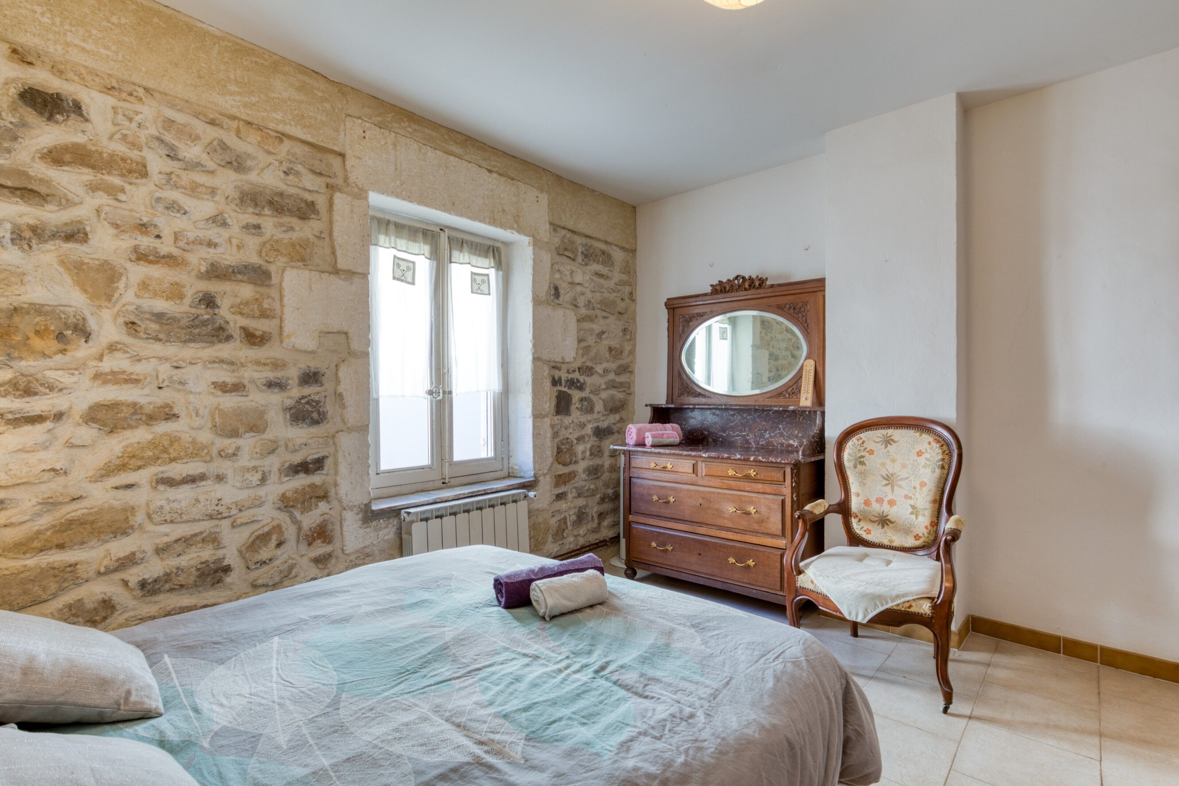 A bedroom with a stone wall, a wooden dresser, a chair, and a bed with a patterned bedspread.