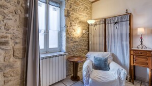 Une pièce aux murs de pierre, un lit recouvert d’une couverture blanche, une petite table ronde, une lampe et une fenêtre avec des rideaux.