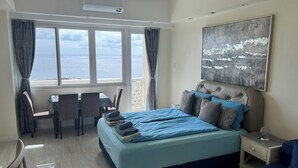 A bedroom with a bed, a desk, and a chair, a bedside table with a lamp, and a large window with curtains. - La Mirada Residences spaces Sea-view Suites (Lapu-Lapu)