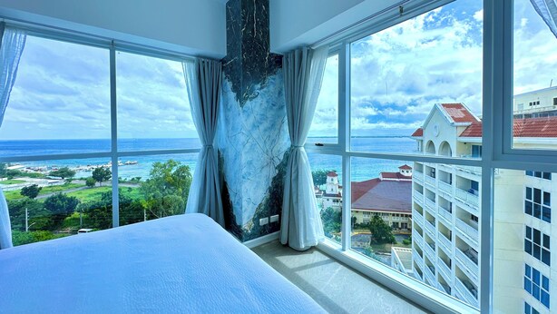 A hotel room with a large bed, a marble accent wall, and a view of the ocean and buildings. - La Mirada Residences spaces Sea-view Suites (Lapu-Lapu)
