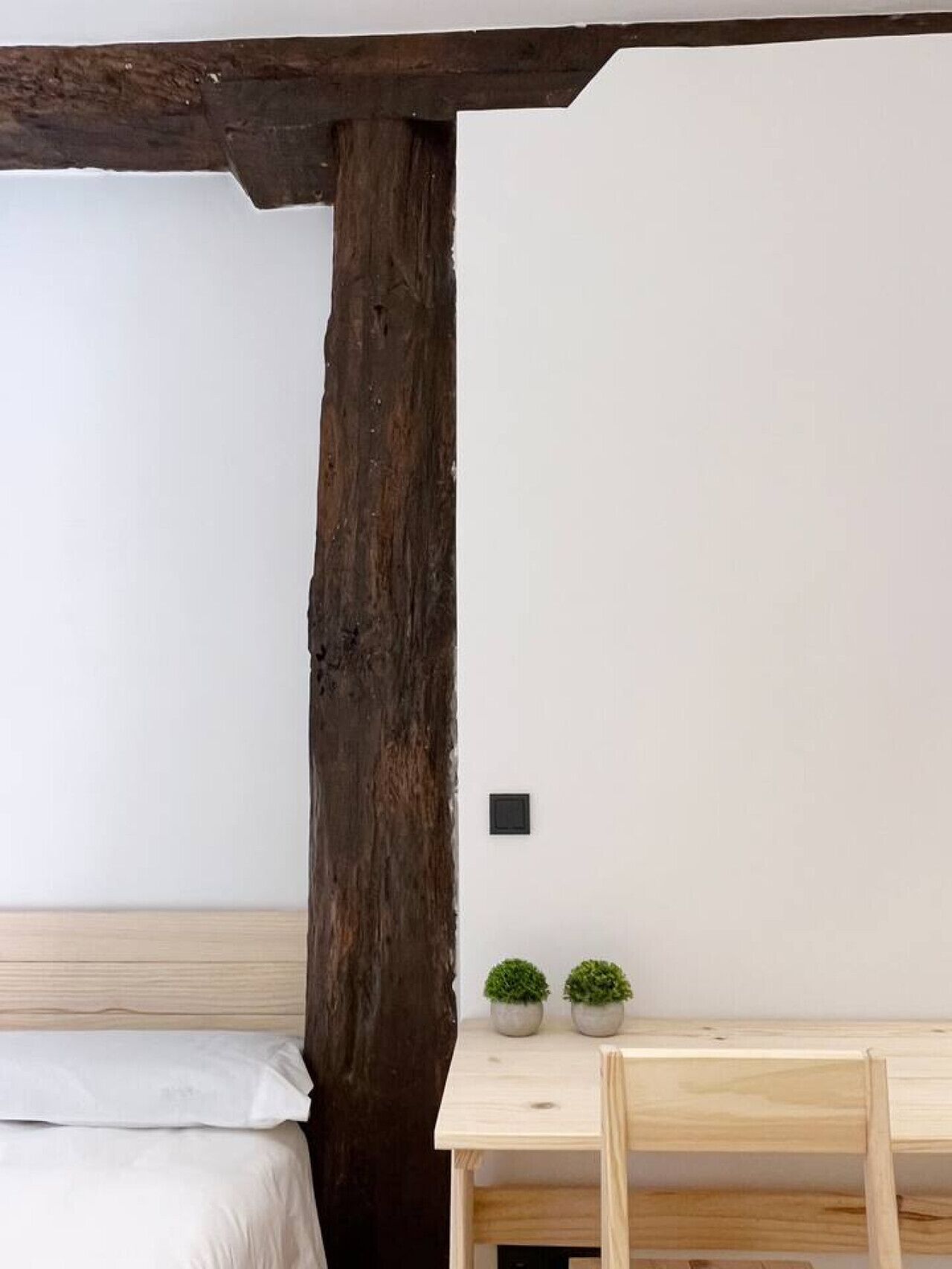Habitación con techo de viga de madera, cama con ropa blanca, una mesa pequeña con dos plantas en macetas y un interruptor de luz fijado a la pared.