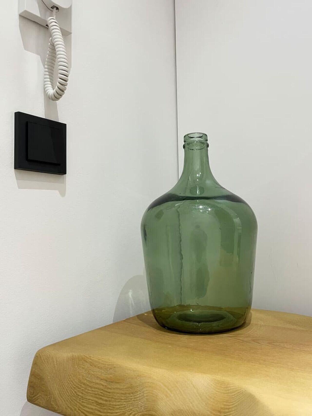 Una botella de vidrio verde sobre una mesa de madera frente a una pared blanca, con un interruptor negro y un teléfono de cable blanco.