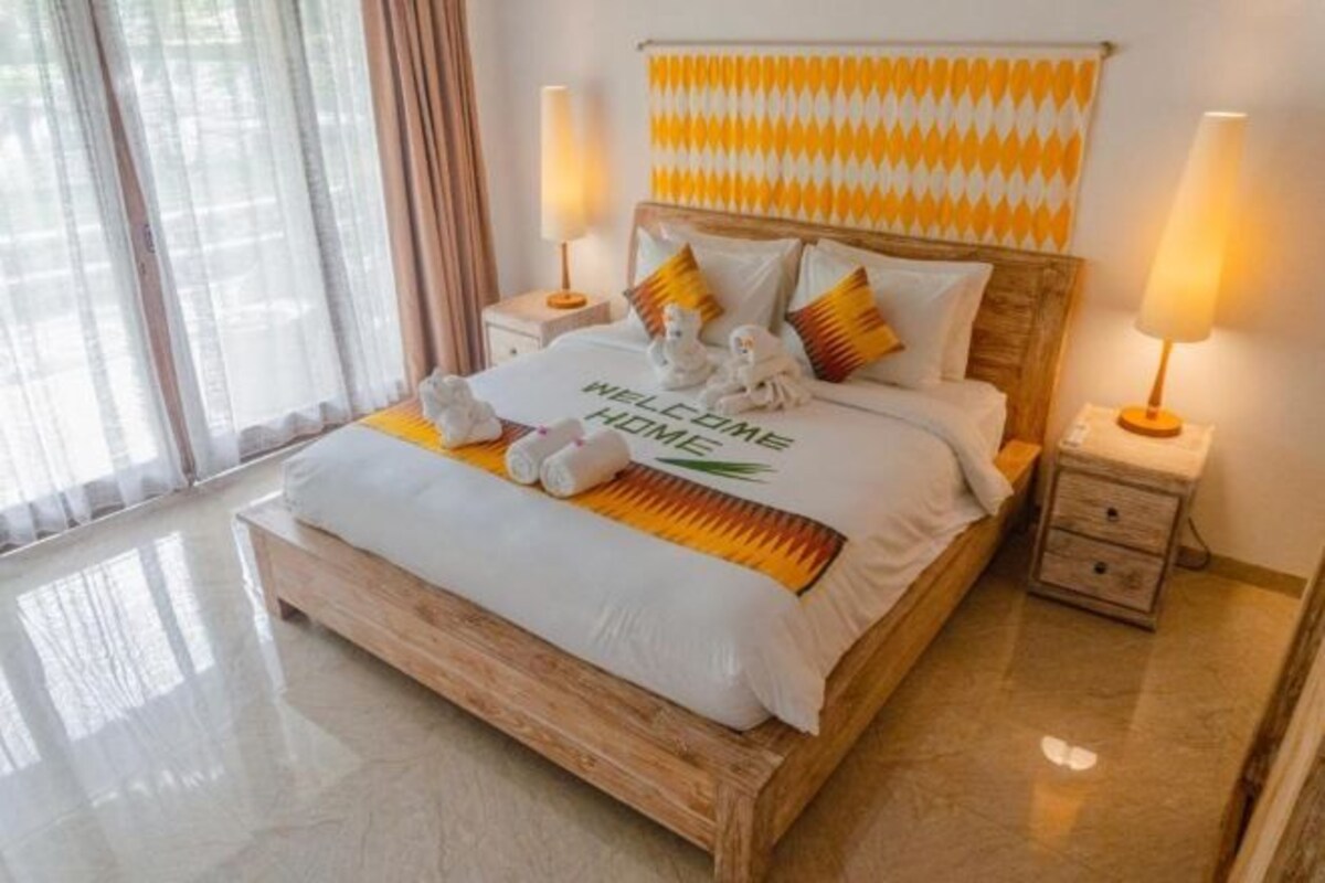A hotel room with a wooden bed, a 'Welcome Home' sign, two bedside lamps, and a view of the outdoors through sheer curtains.