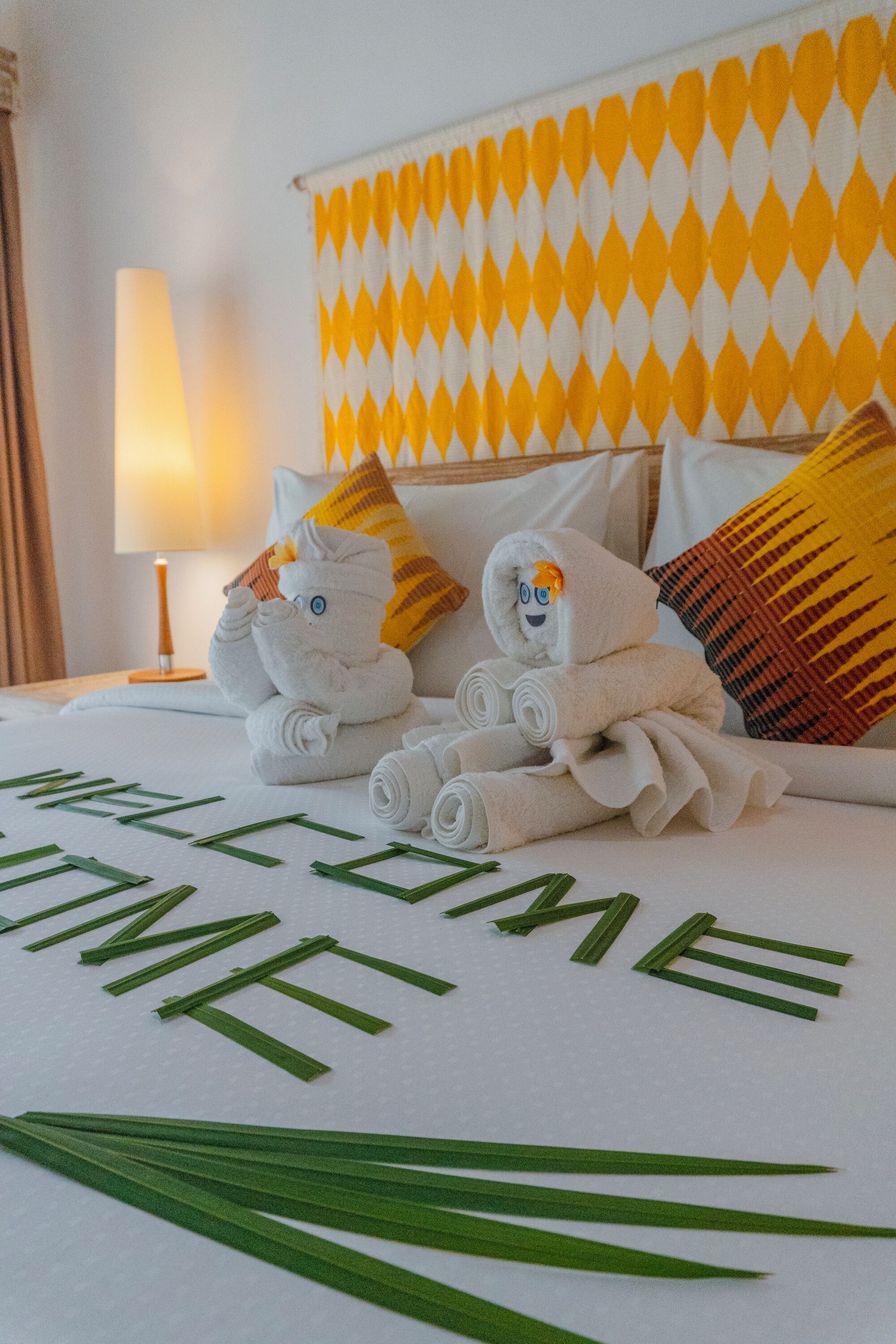 Una cama bien hecha con toallas enrolladas, un peluche, una lámpara y un cabecero estampado.