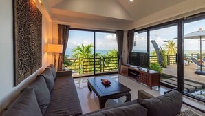 A modern living room with a sofa, a coffee table, and a TV. The room has large windows with a view of palm trees and a balcony.