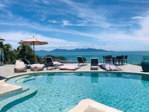 En pool med kristallklart blått vatten, omgiven av solstolar och ett bord med utsikt över havet och bergen.