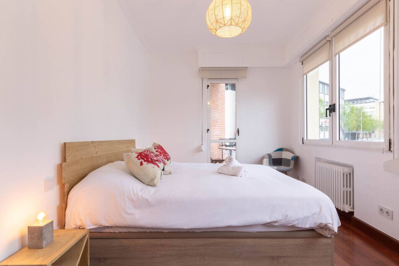 A bedroom with a wooden headboard, white bedding, a bedside table with a lamp, a radiator, and a window with a view of the outside.