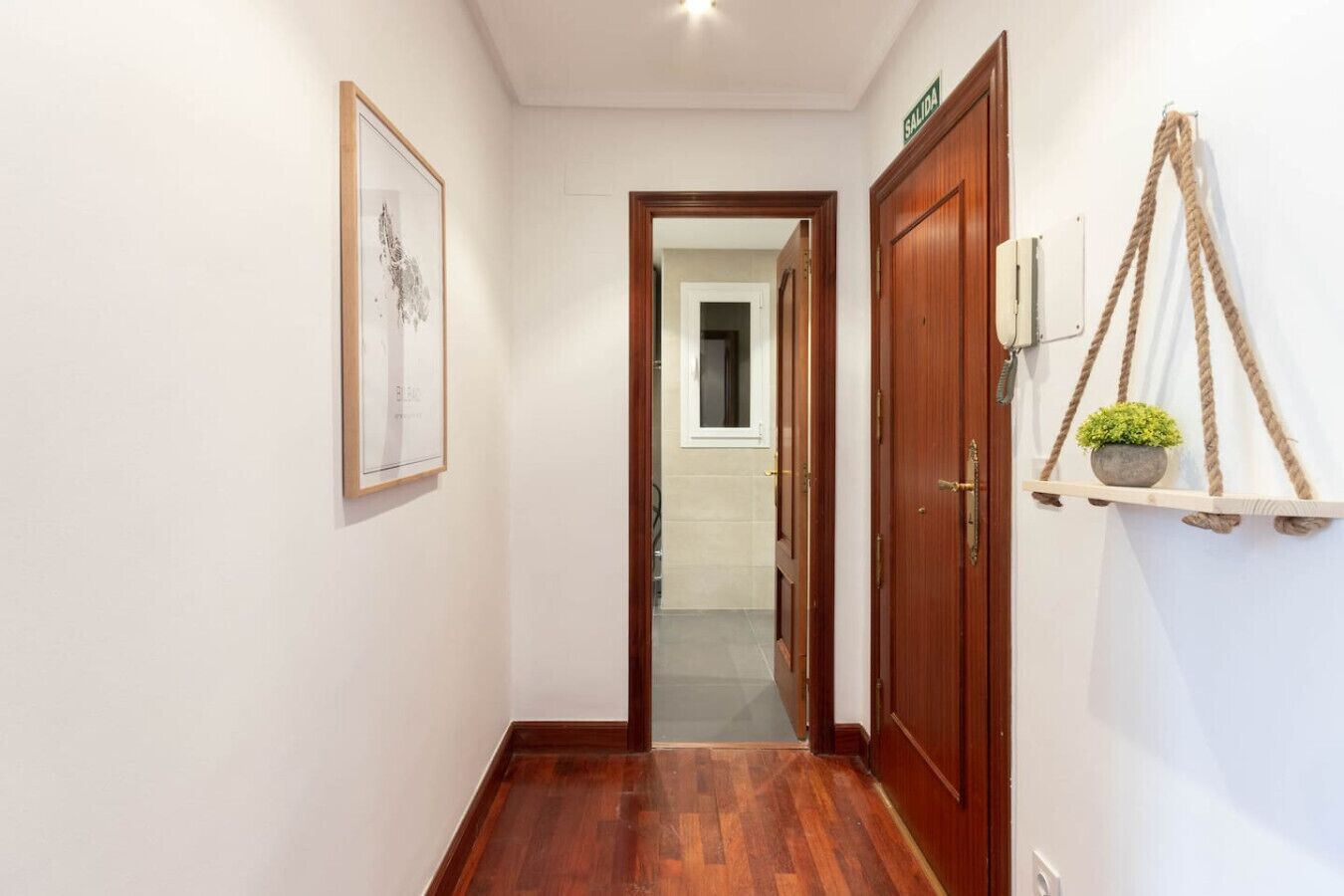 Un couloir avec un plancher en bois, un tableau encadré au mur, une porte avec une pancarte et une plante posée sur une étagère.