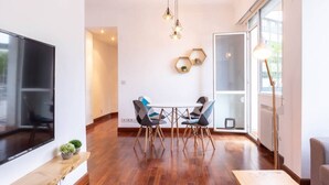 A modern living room with a large TV, wooden flooring, a dining area with a table and chairs, and a large window providing natural light.
