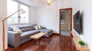 A modern living room with a grey sectional sofa, a wooden coffee table, a floor lamp, a flat-screen TV mounted on the wall, and a wooden cabinet with a plant.