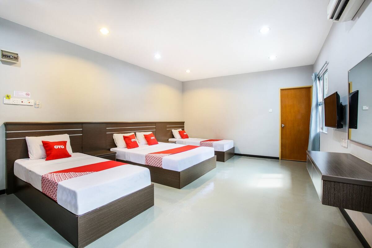 Une chambre d’hôtel avec trois lits, chacun doté de draps blancs et de touches de couleur rouge, d’une tête de lit en bois, d’un téléviseur à écran plat et d’une porte en bois.