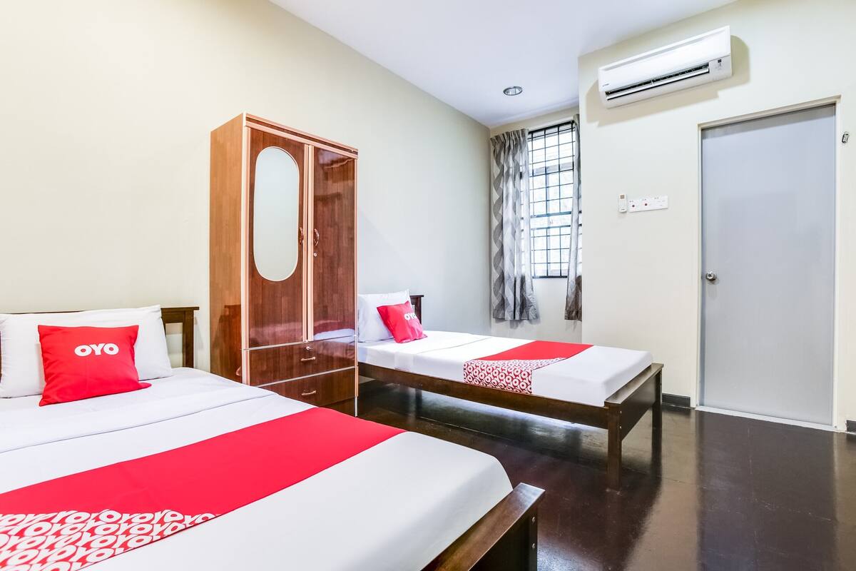 Une chambre d’hôtel avec deux lits, chacun recouvert d’une couverture rouge et blanche, une armoire en bois, une fenêtre avec des rideaux et un appareil de climatisation fixé au mur.