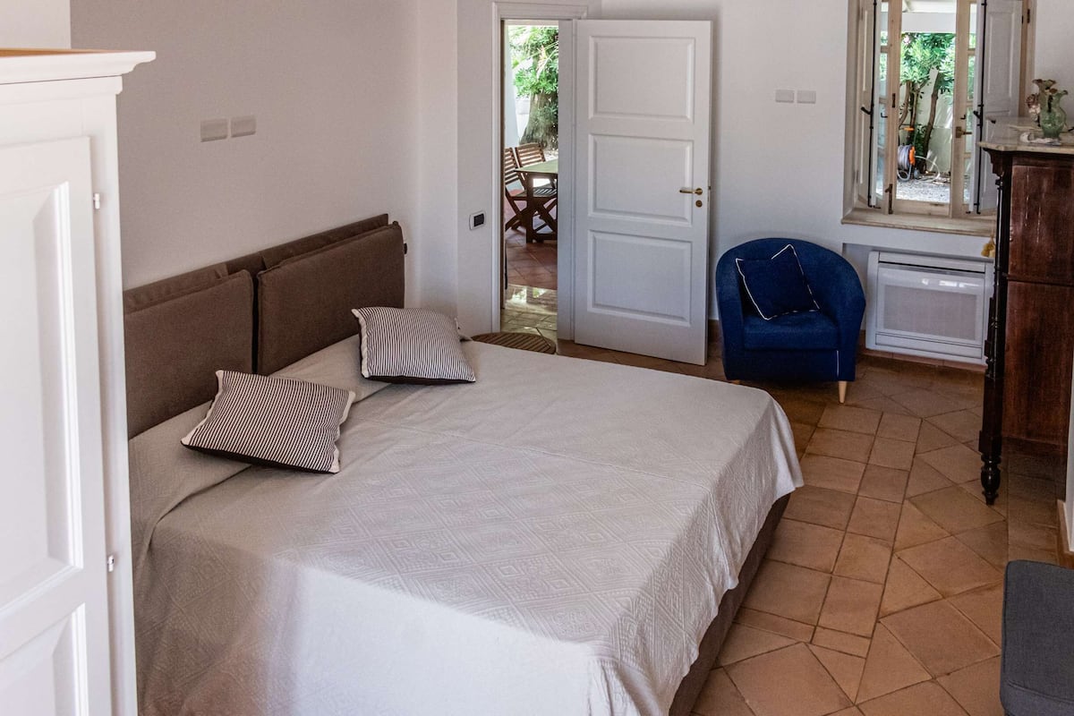 A bedroom with a bed, pillows, a chair, a dresser, and an open door leading to a patio area.