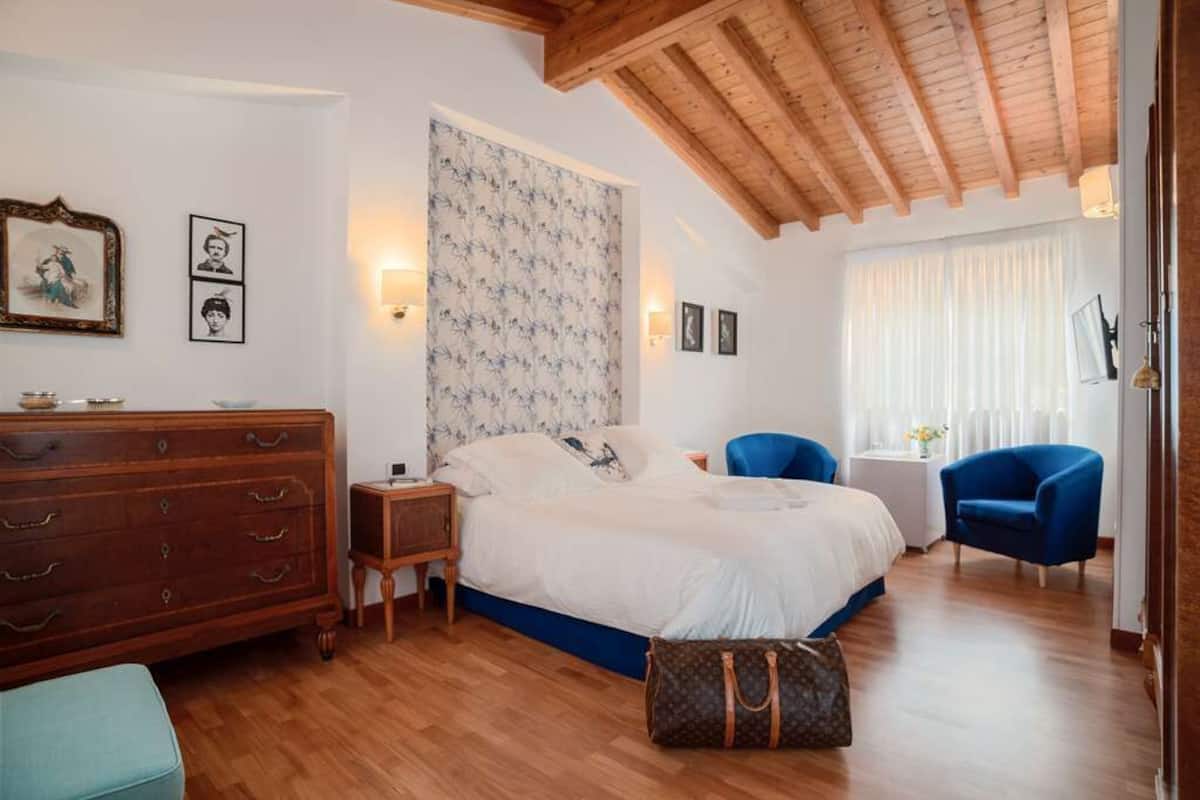 A bedroom with a wooden ceiling, a bed with a floral headboard, a dresser, a blue chair, and a small table with a lamp.