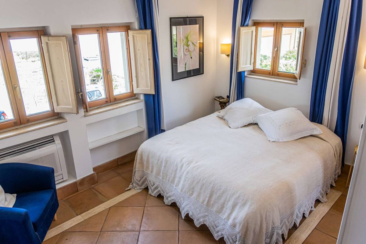 A bedroom with a bed, a chair, a heater, and windows with curtains.