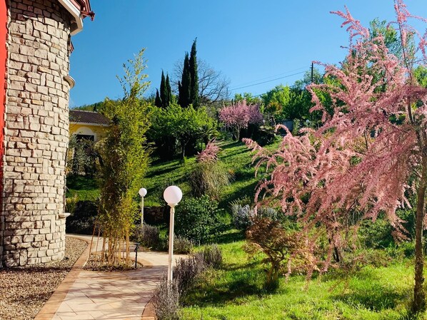 A stone building with a paved pathway leading to a lush garden with various plants and a pink flowering tree. - Villa Vento dei Cieli (Teora)