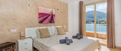 Een slaapkamer met een bed, nachtkastje, koffer, schilderij en een balkon met uitzicht.