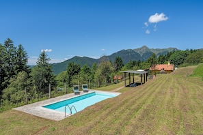 A swimming pool with a stone deck, surrounded by a grassy area, trees, and mountains in the background.
