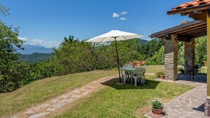 Eine Terrasse mit Tisch und Stühlen, einem weißen Sonnenschirm, Steinpfeilern und Blick auf Berge.