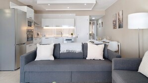 Ein modernes Wohnzimmer mit einem grauen Sofa, einem Esstisch mit Stühlen, einer Küche mit weißen Schränken und einem großen Fenster.
