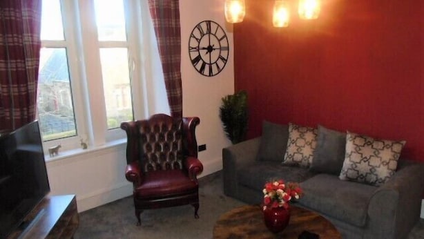 A living room with a red wall, a clock, a sofa, a chair, and a coffee table with a vase. - Snug - Abhainn Apartment (Helensburgh)