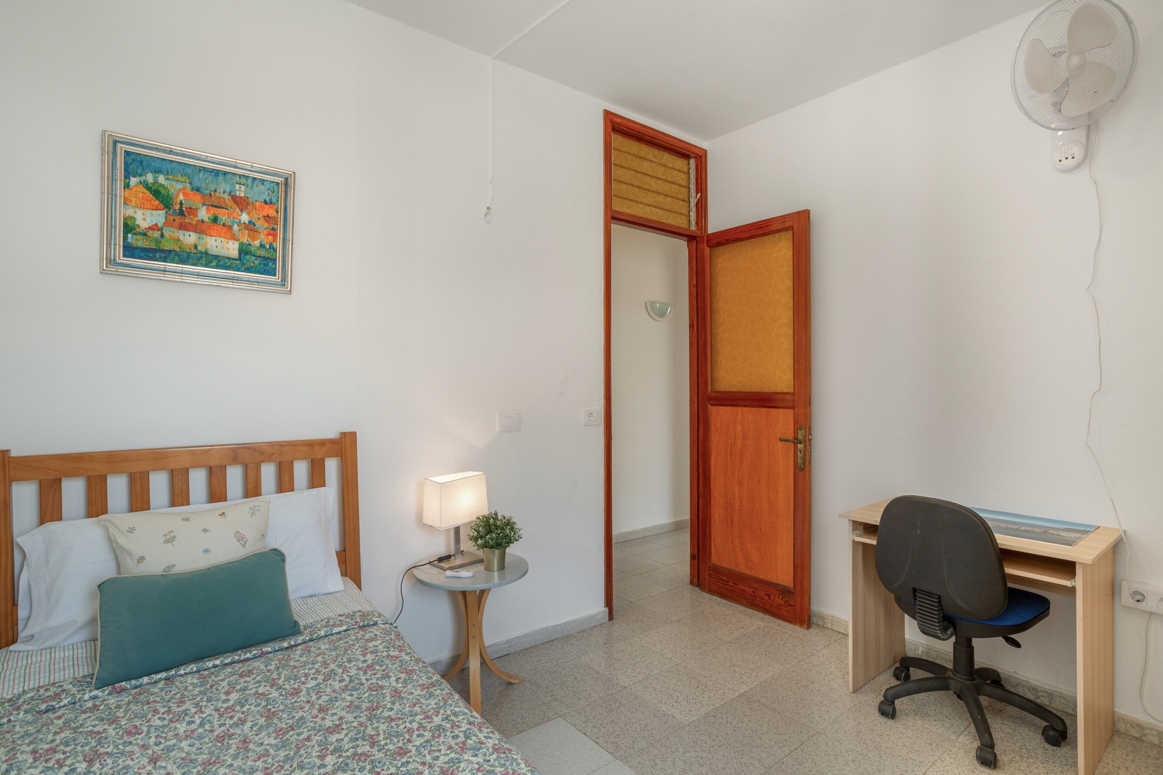 Een slaapkamer met een bed, nachtkastje met lamp, bureau met stoel en een schilderij aan de muur.