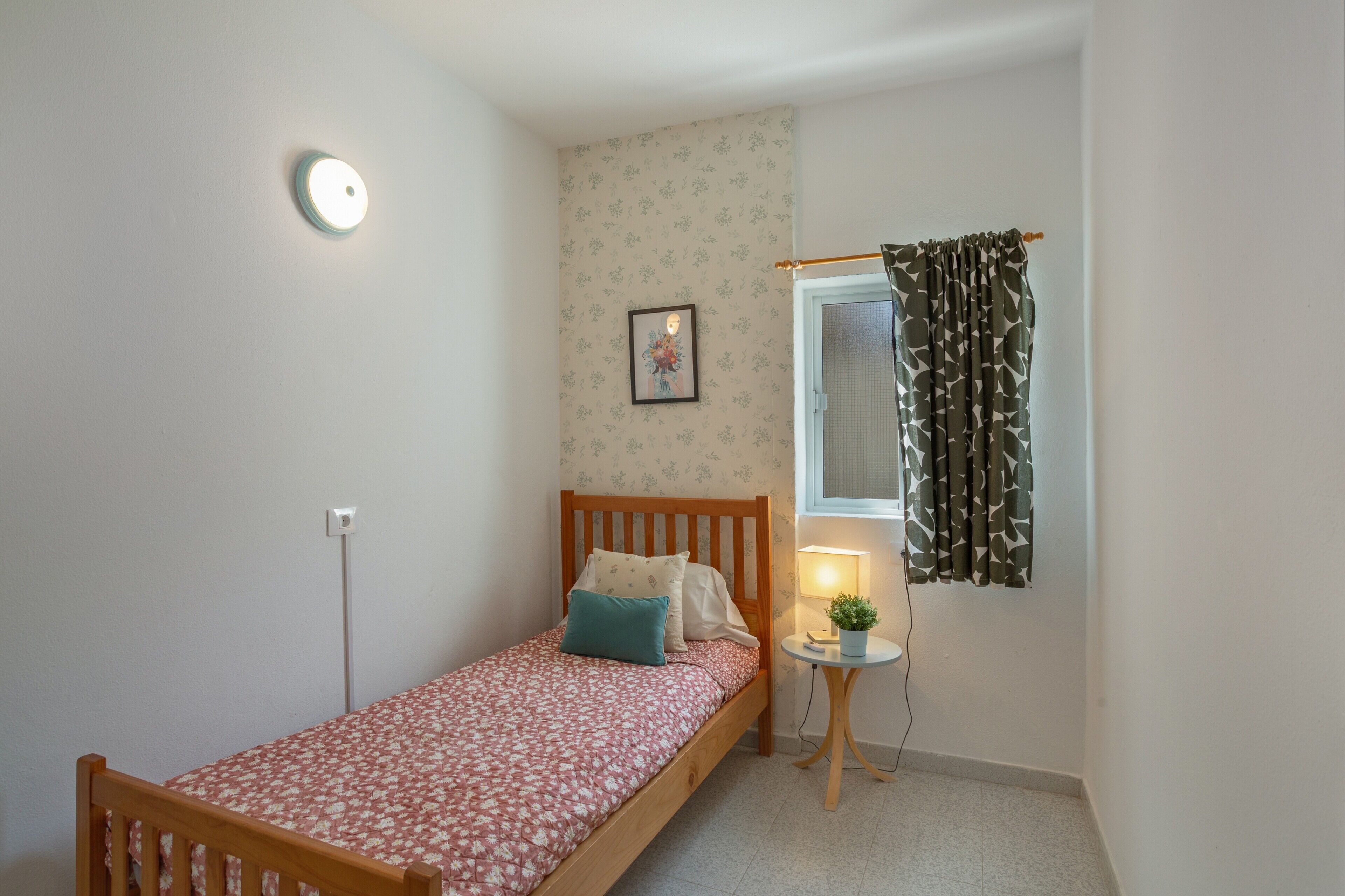 Een eenpersoonsbed met een bloemenbedekking, een bedlampje, een klein tafeltje met een plant, een ingelijst beeld aan de muur en een raam met geruit gordijnen.
