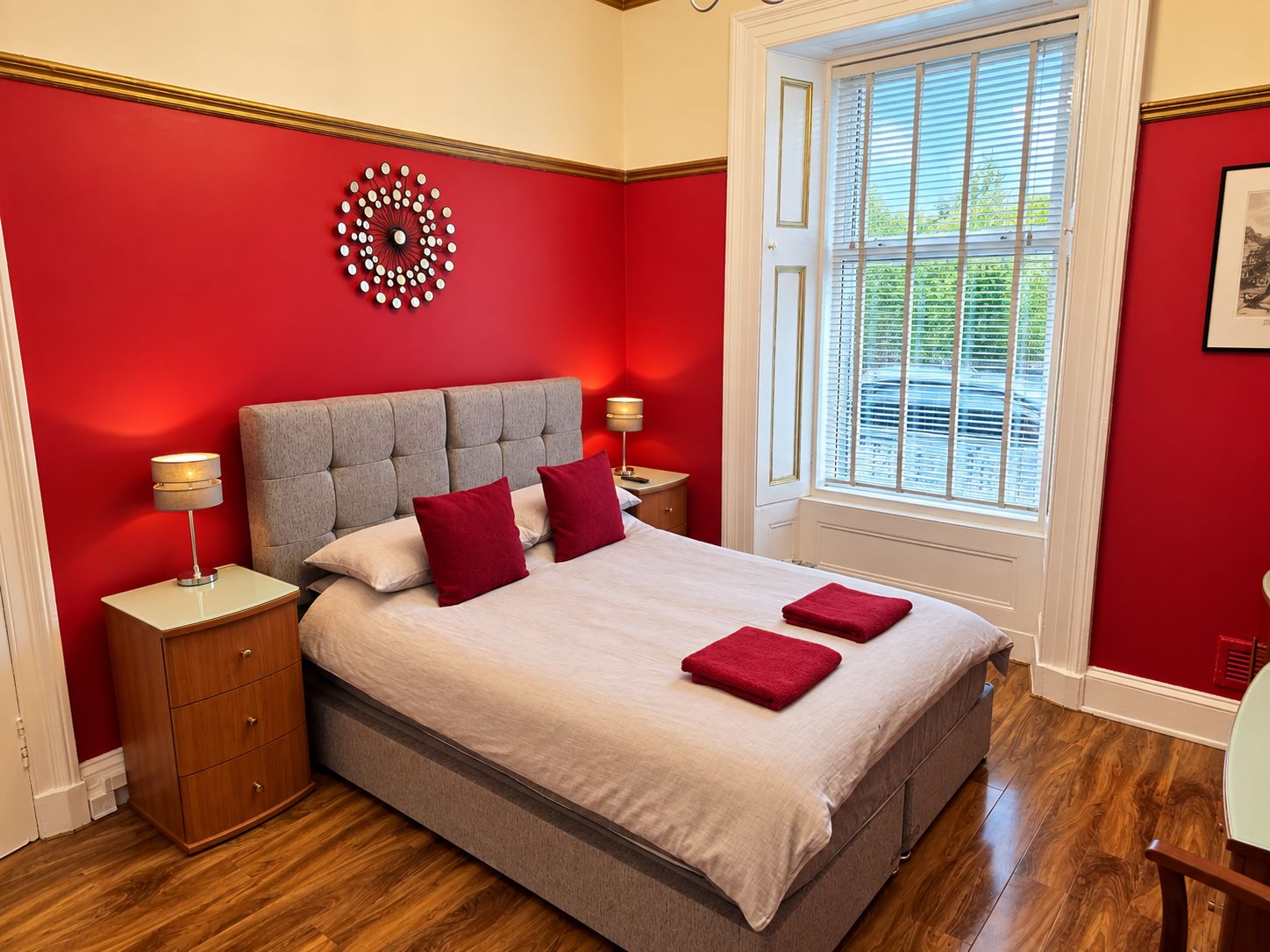 A bedroom with a large bed, two nightstands, a red wall, a window with blinds, and a decorative wall piece.