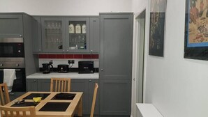 Eine kompakte Küche mit grauen Schränken, einer Mikrowelle, einem Wasserkocher und einem Brotbackgerät. Ein Esstisch mit Stühlen und ein kleines Fenster sind ebenfalls vorhanden.