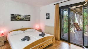 Une chambre avec un lit en bois, deux lampes de chevet, un radiateur et une porte-fenêtre coulissante donnant sur un espace extérieur aménagé avec une chaise et des plantes.