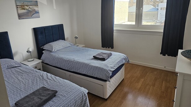 A bedroom with two beds, a bedside table with a lamp, a window with curtains, and a painting on the wall. - Snug - Colquhoun Square Apartment (Helensburgh)