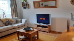 A living room with a sofa, a coffee table, a TV on a wooden stand, and a mirror on the wall. - Snug - Colquhoun Square Apartment (Helensburgh)