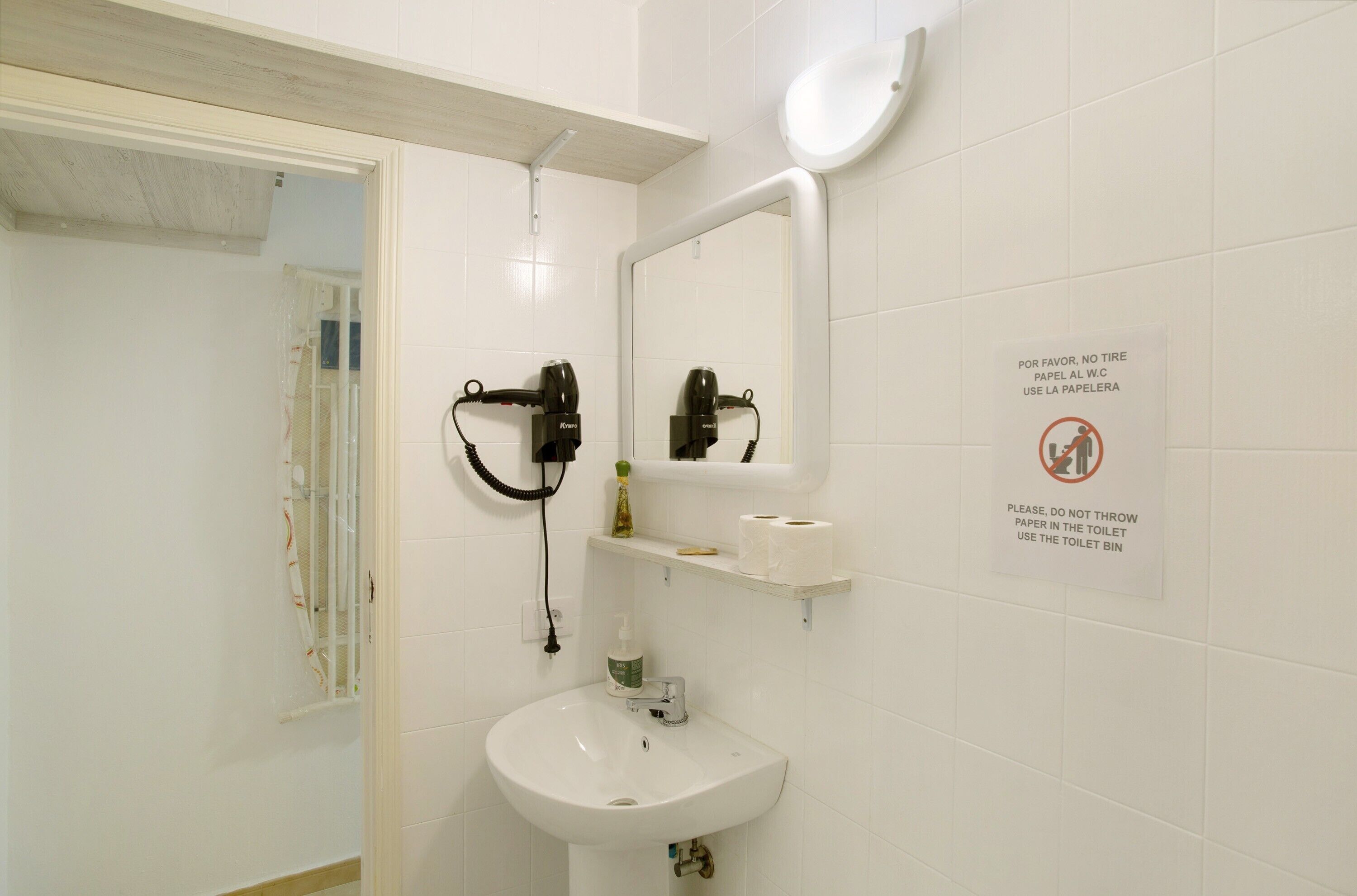 Een badkamer met een wastafel, spiegel en twee haardroger. Er staan bordjes met instructies om geen papier door het toilet te spoelen en de prullenbak te gebruiken.