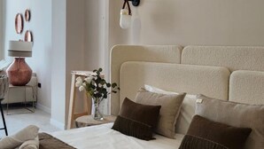 Hotellihuone, jossa on sänky, tyynyjä, yöpöytä lampun kanssa, maljakko kukkineen ja seinäkello.