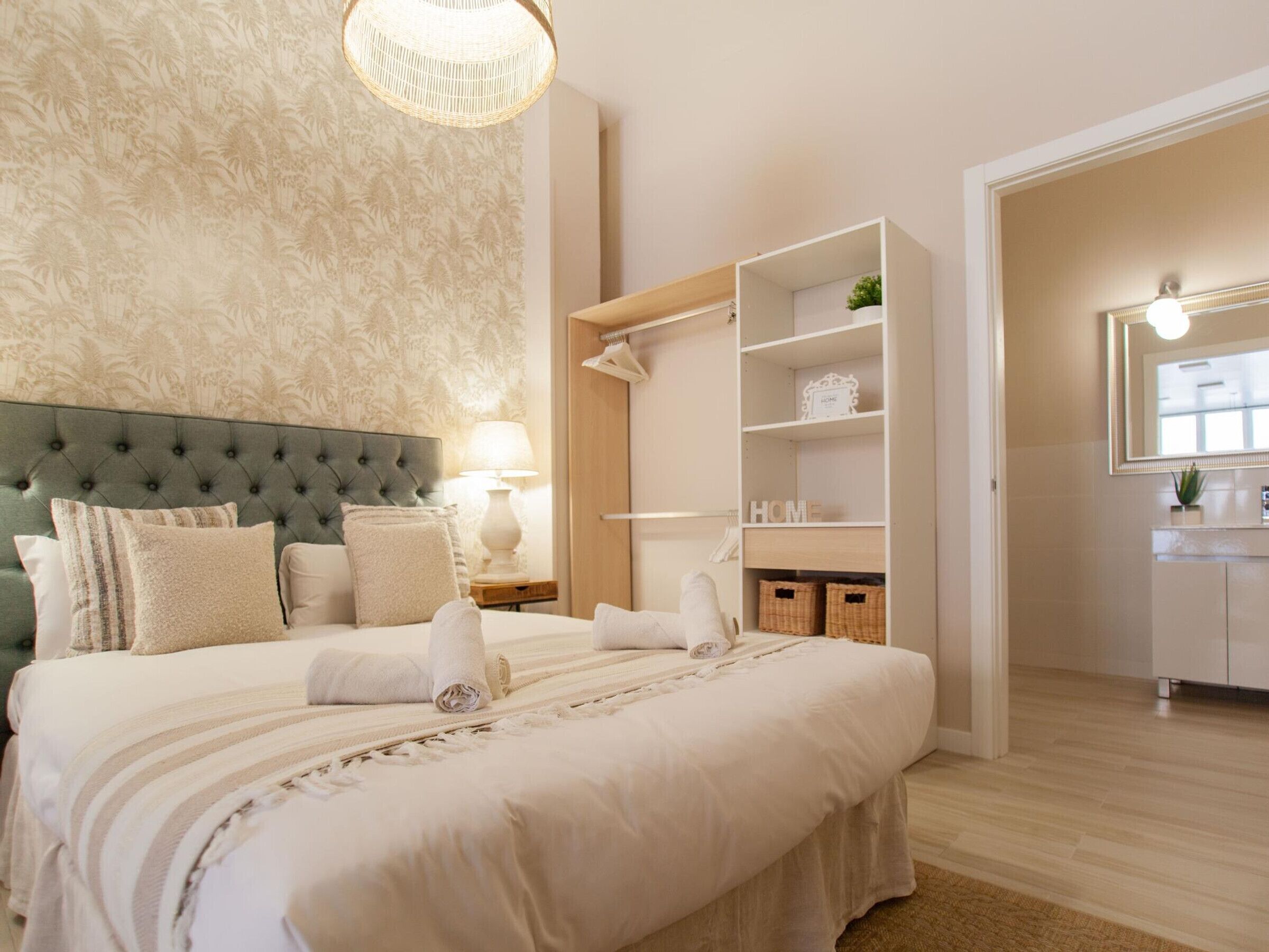 Un dormitorio ordenado con cabecera capitonada, una cama grande con toallas enrolladas, una lámpara de noche y una unidad de estanterías empotradas con cestas.