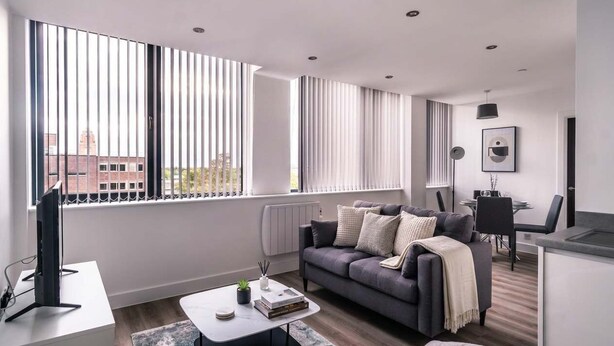 Una sala de estar moderna con un sofá, una mesa de centro y un televisor. Hay ventanas grandes con persianas, un radiador y un comedor con sillas.