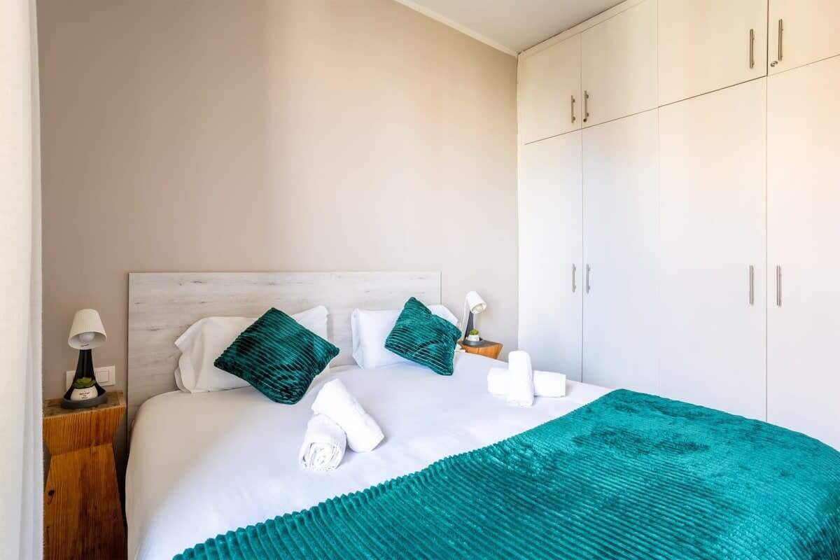 A hotel room with a bed, white curtains, a wooden nightstand, a teal throw blanket, and a white headboard.