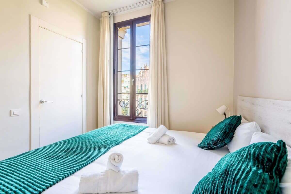 A hotel room with a bed, a window with curtains, a view of buildings outside, and a door.