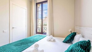 Ein Hotelzimmer mit Bett, einem fenster mit Vorhängen, Blick auf Gebäude und einer Tür.